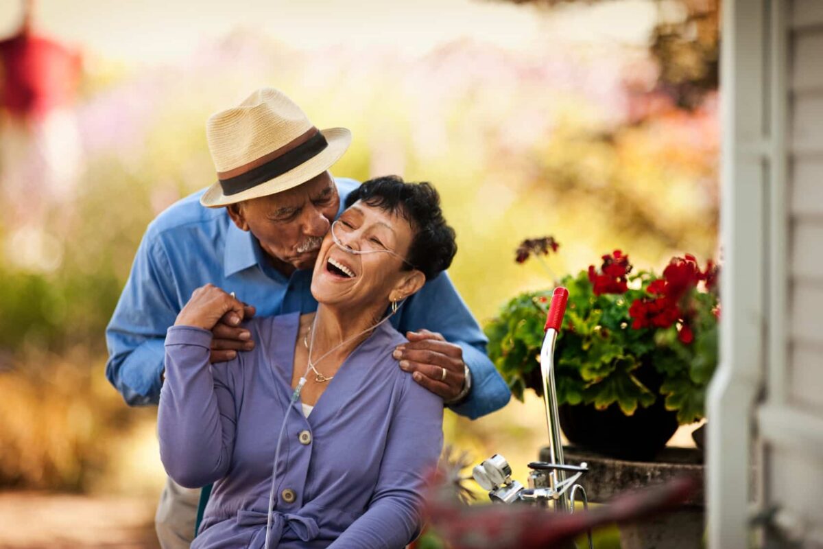 A man kisses his wife from behind who sits with her portable oxygen concentrator machine