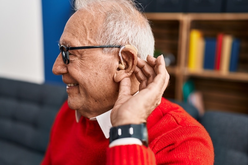 older adult in red sweater models hearing aid in ear