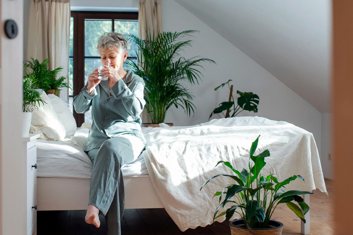 Older adult sitting on edge of a bed drinking a glass of water
