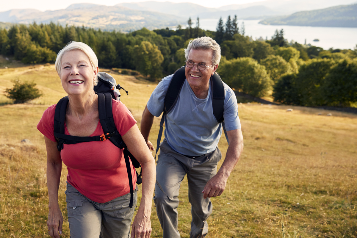 Older adult man and woman hiking up a grassy hill with woods and river in the background