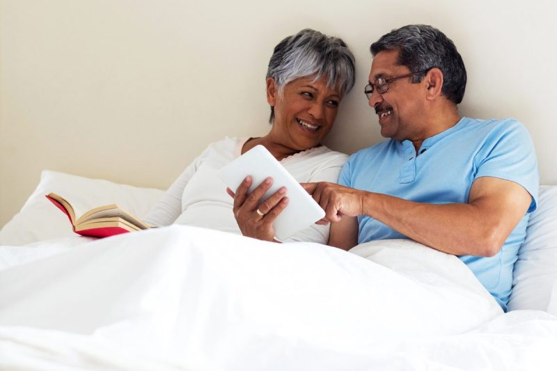 Smiling woman and man relax in bed with white sheets, with man showing the woman something on his tablet while woman reads a book