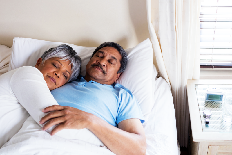 A man and woman lie in bed together with their eyes closed