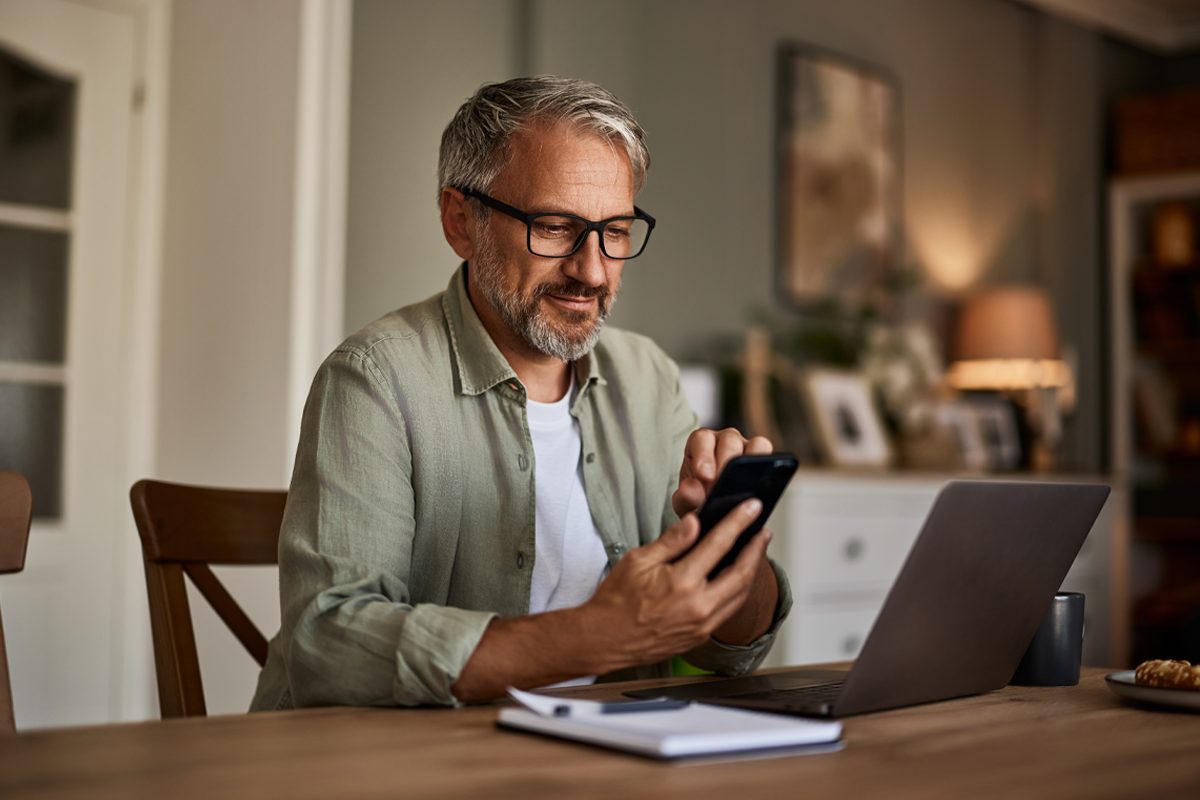 A man wearing glasses sitting at a table looking at a smartphone.