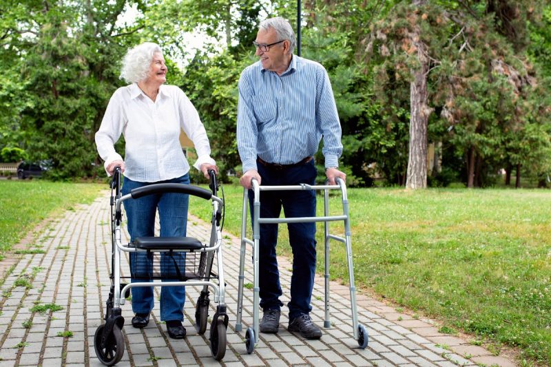 Man and woman smile as they walk outside using their walkers