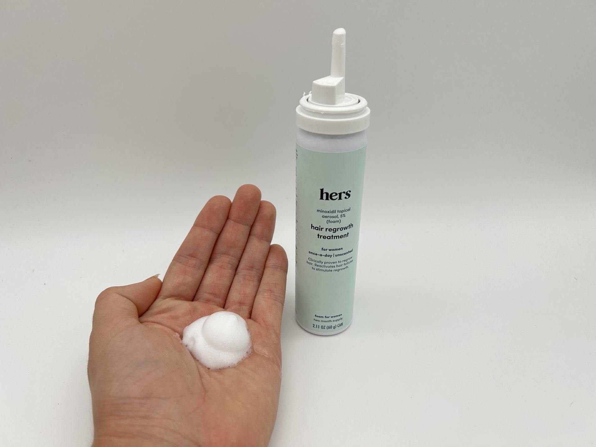 A person’s hand holding a spray of Hers Minoxidil Foam next to a bottle of the foam.